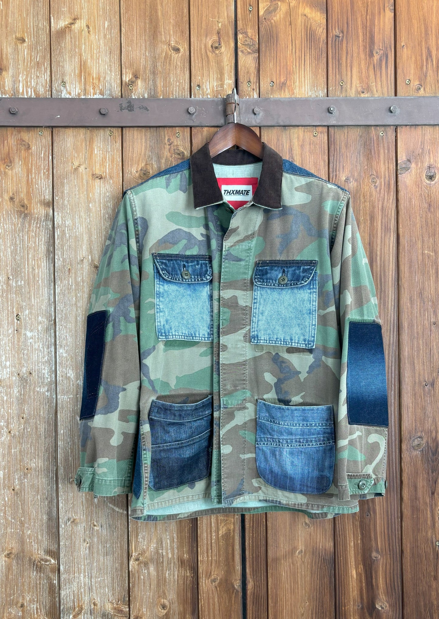 Camouflage-Jacke mit vier aufgesetzten Jeans-Taschen und verstärkten Ellenbogen- und Schulterpartien aus Denim. Der Kentkragen ist aus dunkelbraunem Cordstoff gefertigt und die Jacke wird mit einem Knopfverschluss geschlossen.