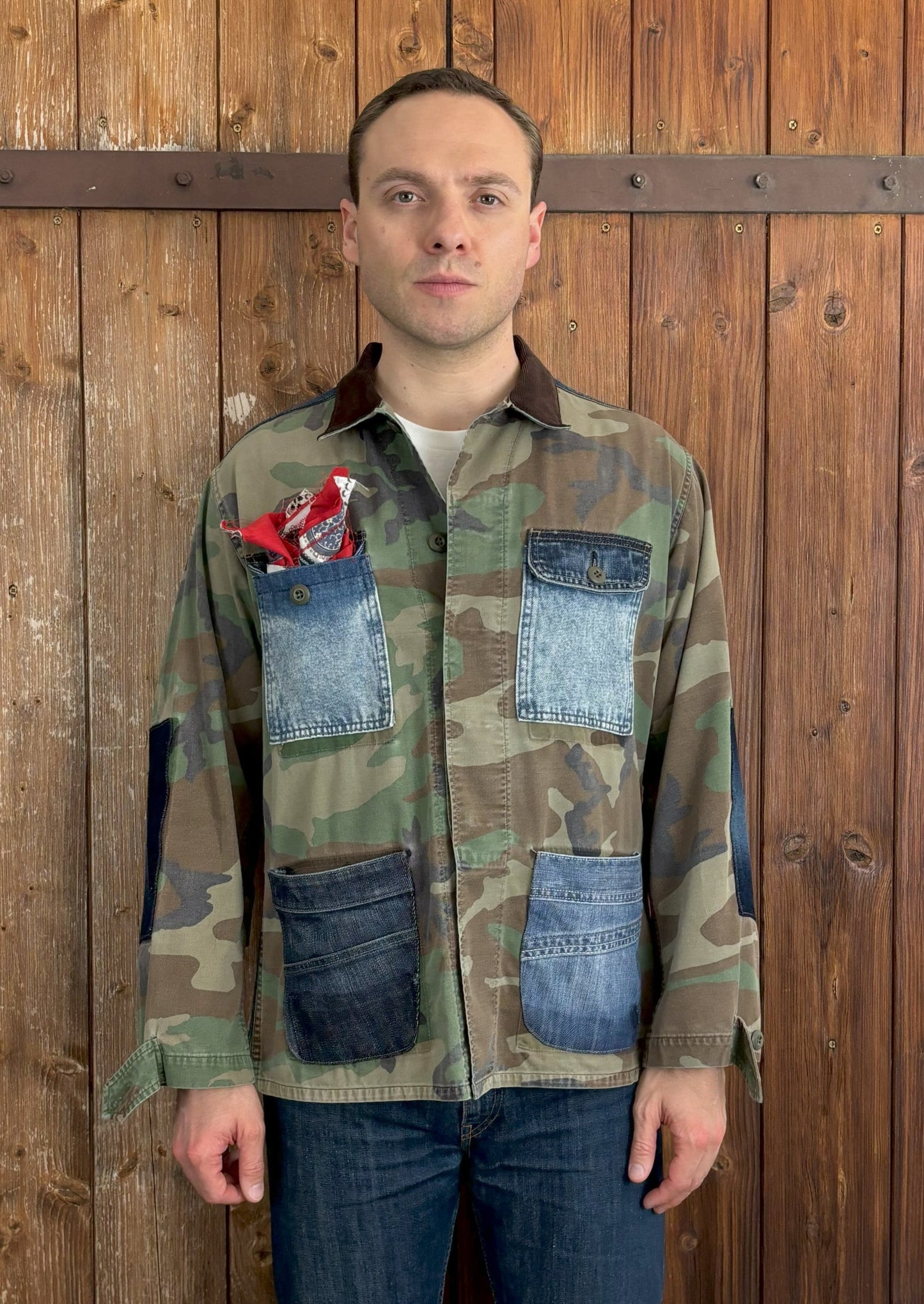 Camouflage-Jacke mit vier aufgesetzten Jeans-Taschen und verstärkten Ellenbogen- und Schulterpartien aus Denim. Der Kentkragen ist aus dunkelbraunem Cordstoff gefertigt und die Jacke wird mit einem Knopfverschluss geschlossen. In der linken Brusttasche befindet sich ein rot-weißes Einstecktuch.